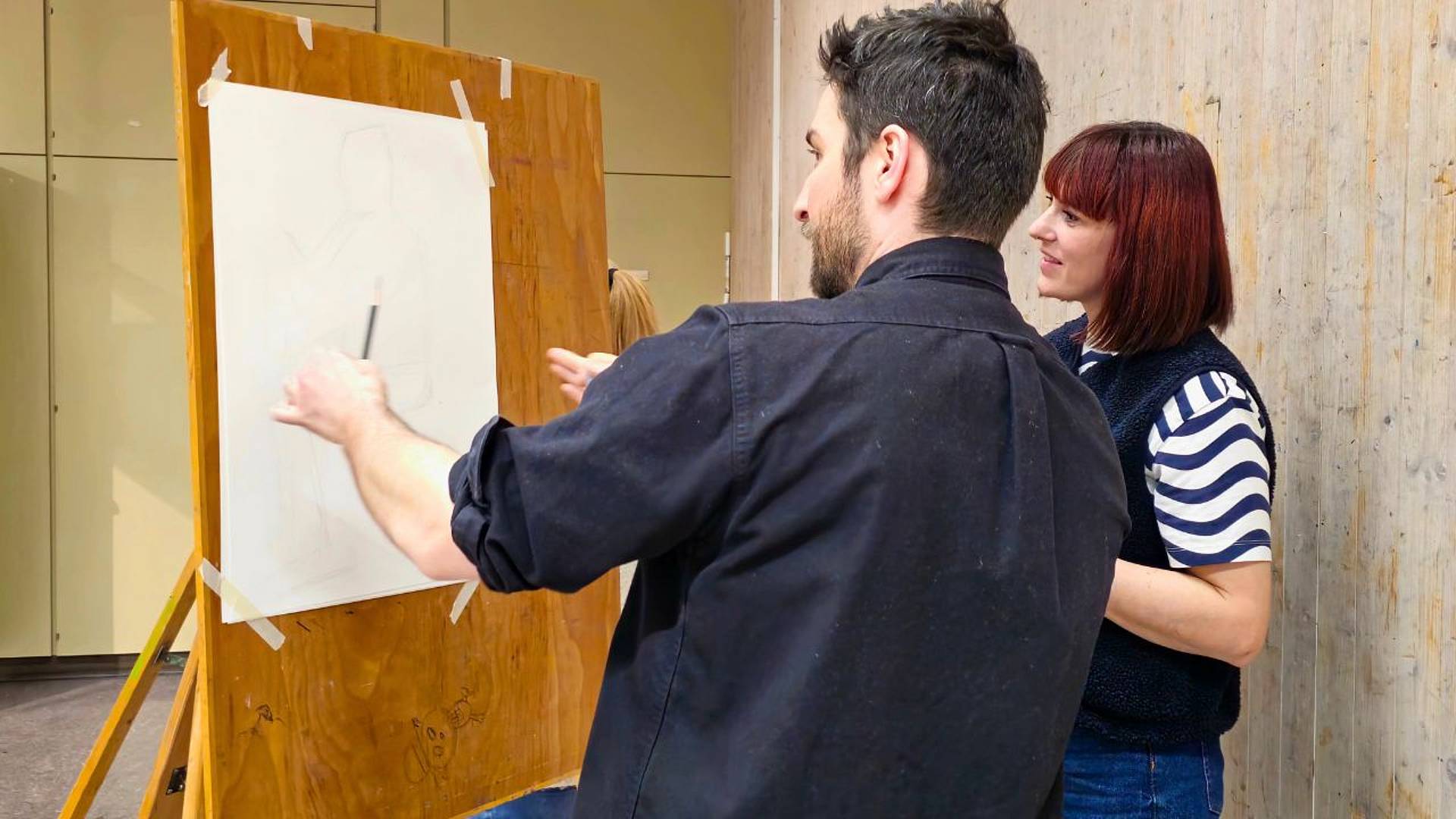 Zwei Personen stehen vor einer Staffelei und besprechen eine entstehende Figurenzeichnung auf Papier.