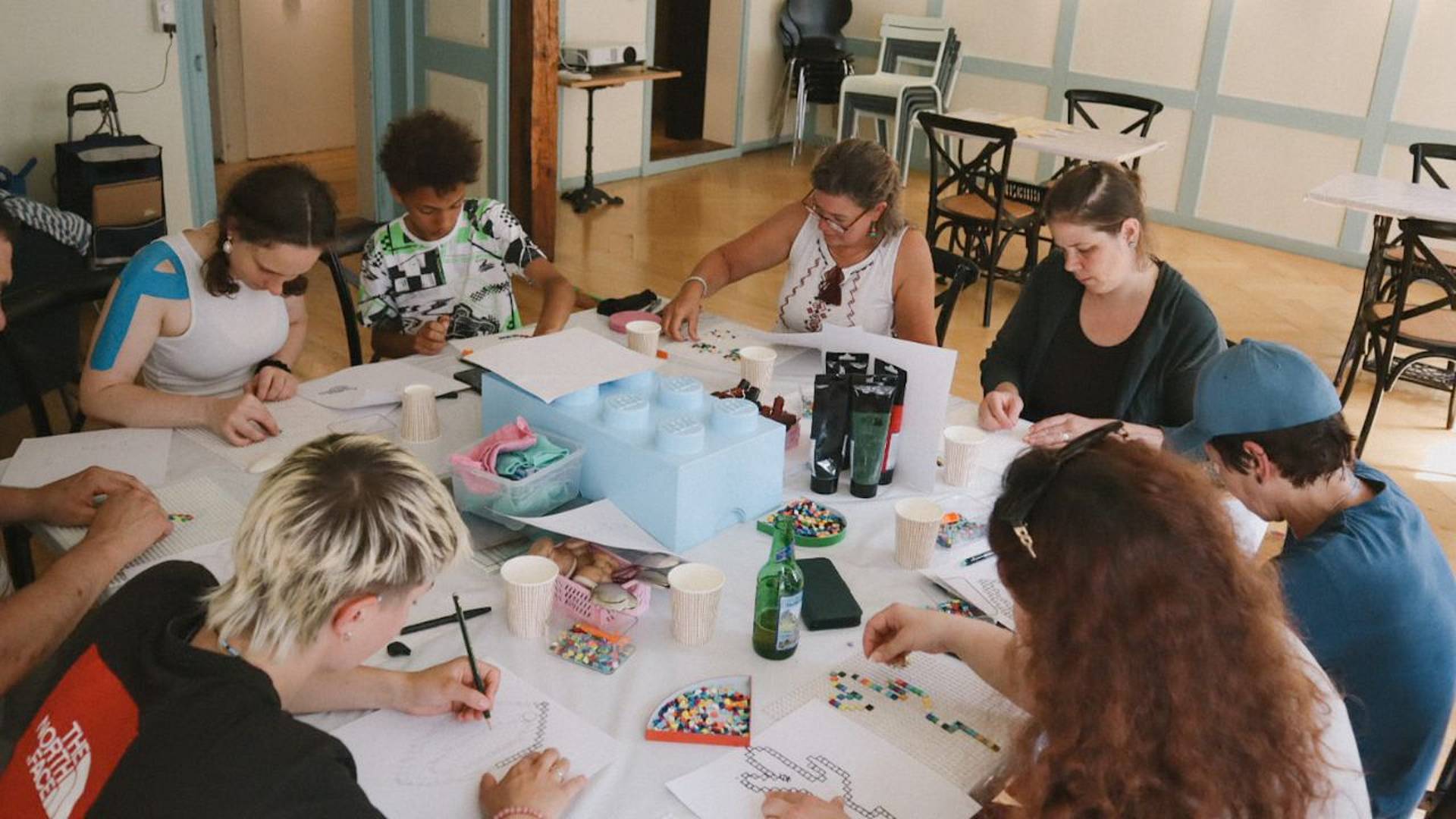 Mehrere Personen arbeiten an einem Tisch mit Lego‑Steinen und Papier an Druckmotiven während eines kreativen Workshops.