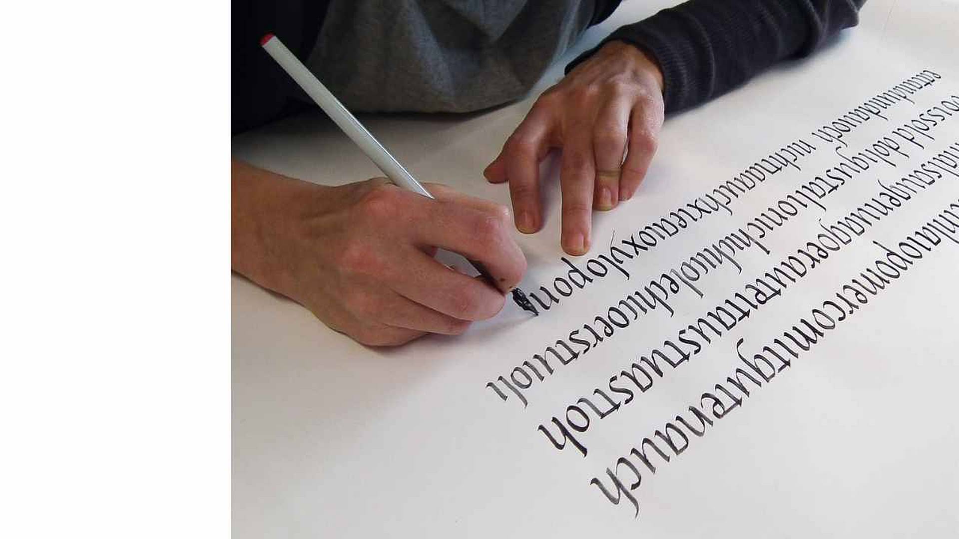 Hände schreiben mit einem schwarzen Kalligrafiestift dekorative Buchstaben auf ein weisses Blatt Papier.