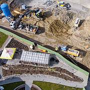 Luftaufnahme einer Baustelle mit Baggern, LKW und Baugeräten neben einem modern gestalteten Außenbereich mit Wegen und Grünflächen.