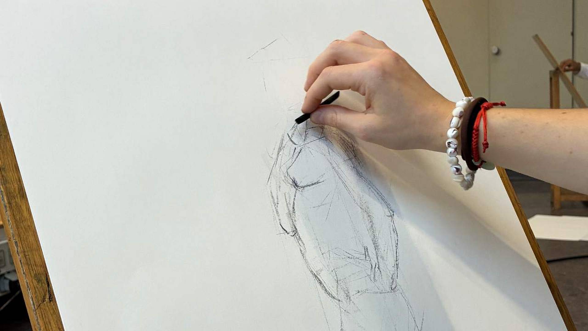 Hand zeichnet mit Kohlestift eine Figurenskizze auf weisses Papier, befestigt auf einer Staffelei.