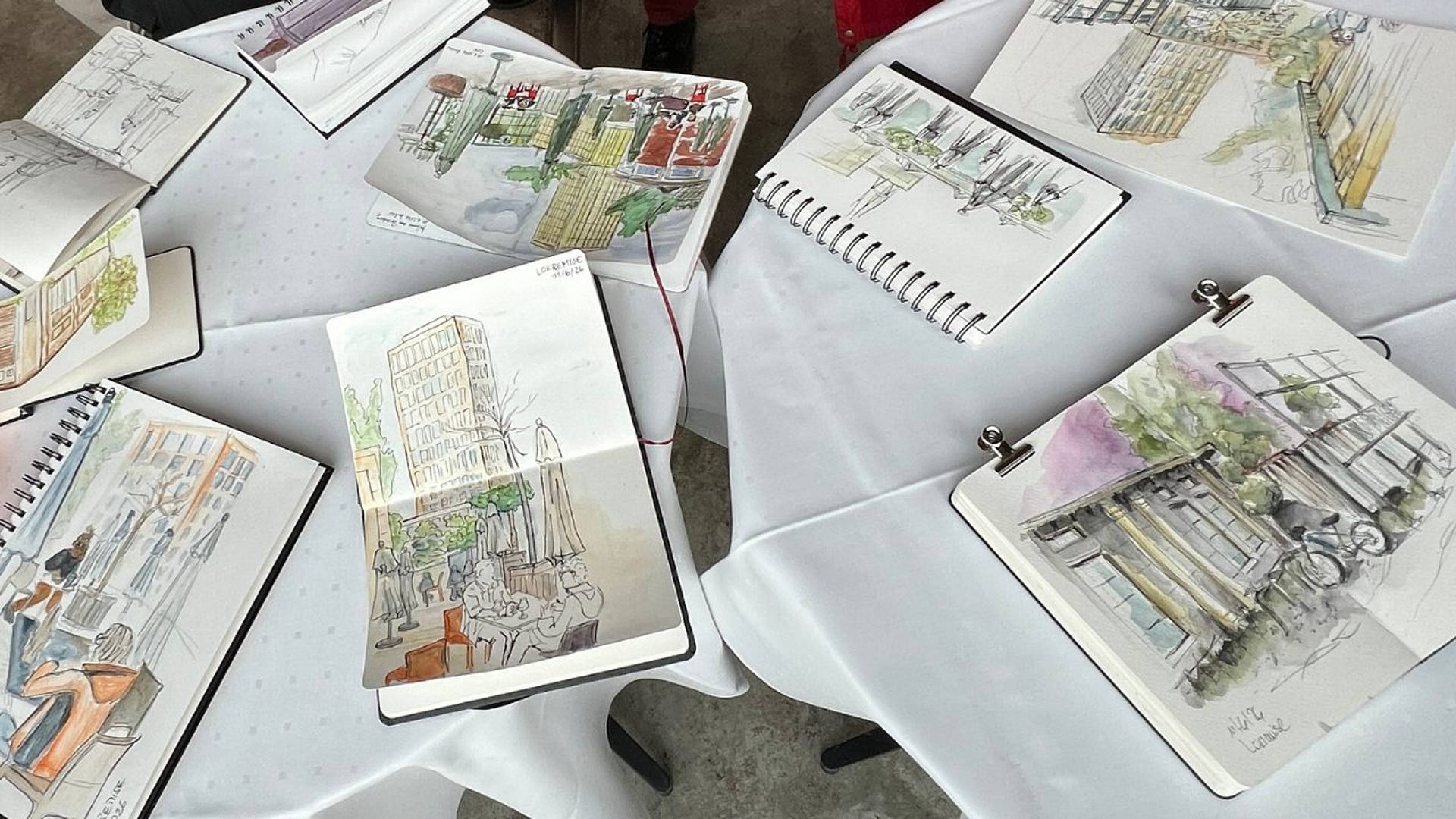 Verschiedene Skizzenbücher mit Aquarellzeichnungen von Stadtansichten, Gebäuden und Cafés liegen offen auf einem Tisch und zeigen unterschiedliche Perspektiven und Details.