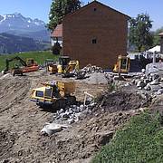 Baustelle im ländlichen Raum mit mehreren Baggern und Erdarbeiten vor alpiner Bergkulisse.