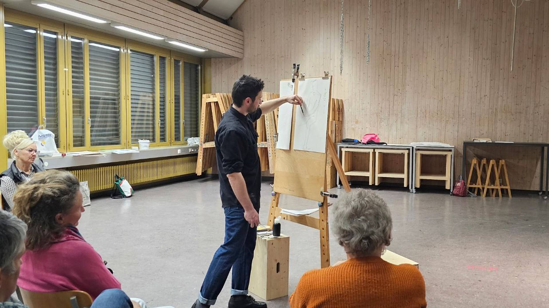 Dozent erklärt eine Figurenzeichnung an der Staffelei, Teilnehmende sitzen und hören zu.