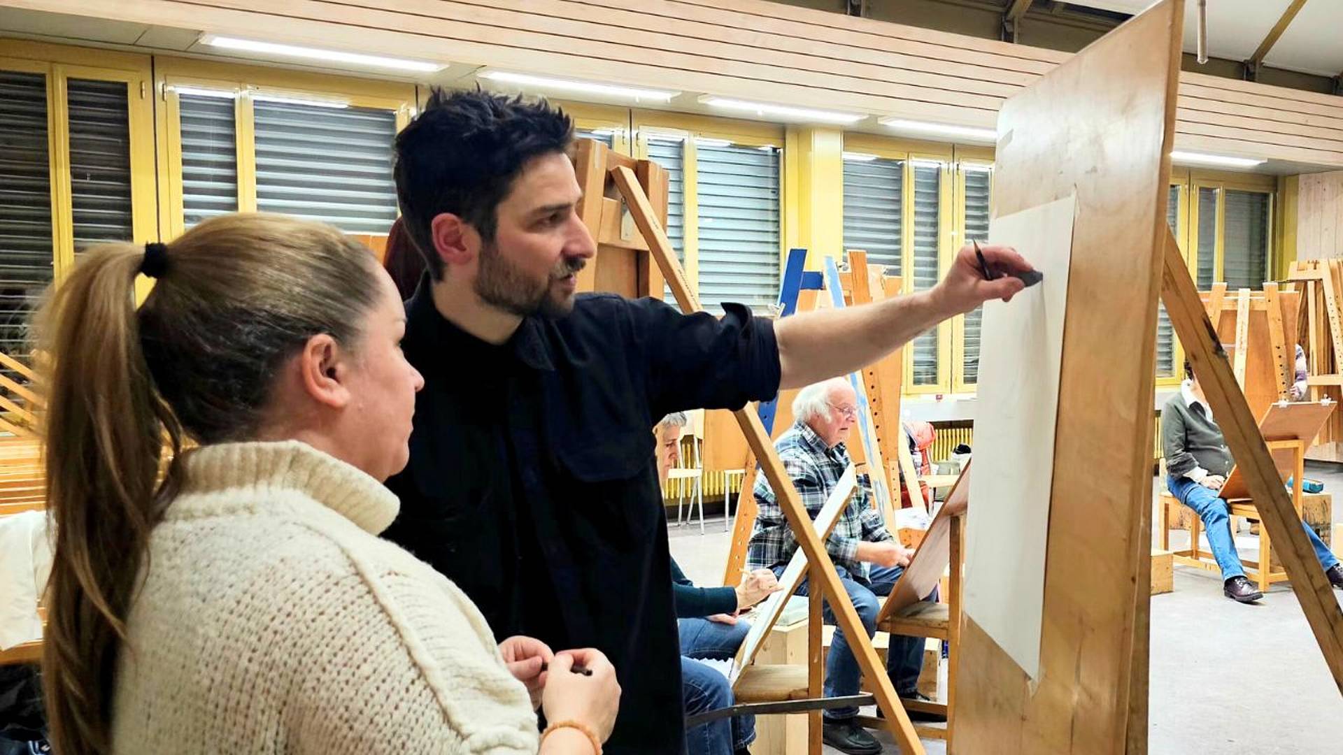 Zwei Personen stehen vor einer Staffelei und besprechen eine entstehende Figurenzeichnung auf Papier.