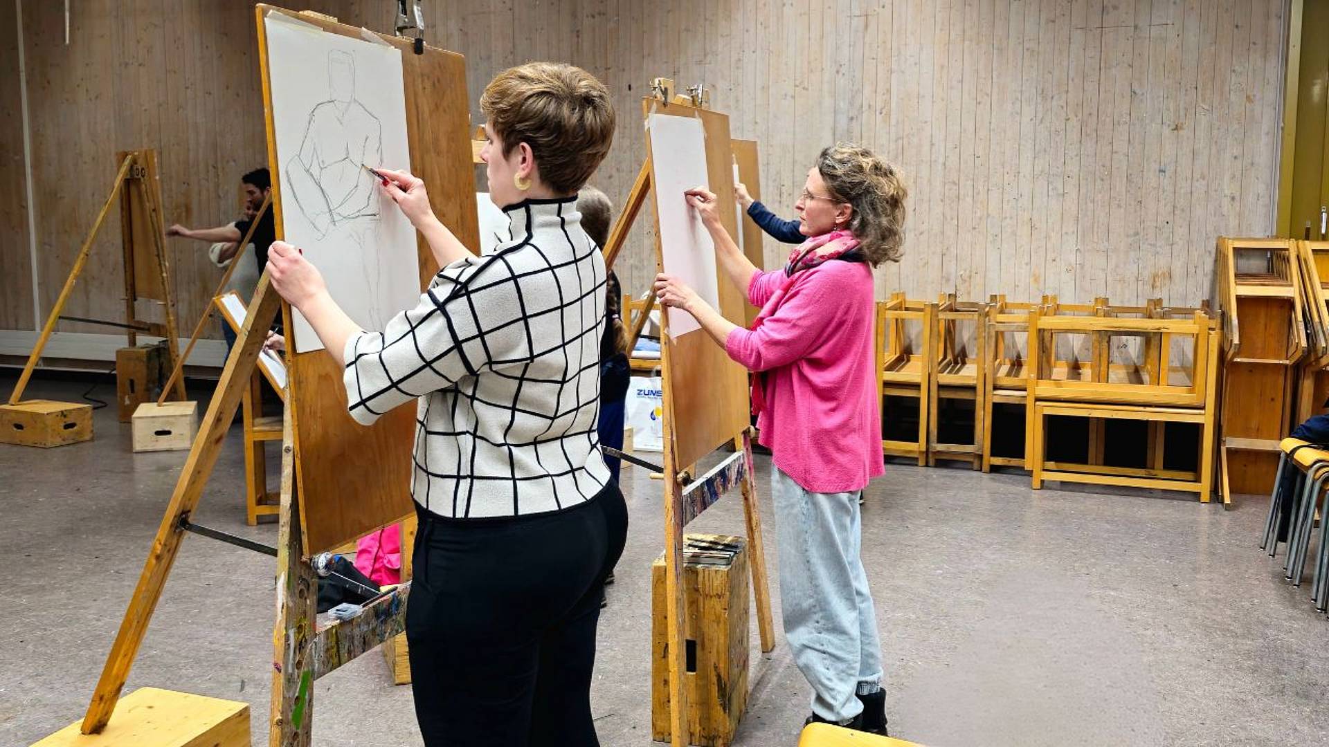 Zwei Personen zeichnen gleichzeitig an nebeneinanderstehenden Staffeleien Figurenstudien.