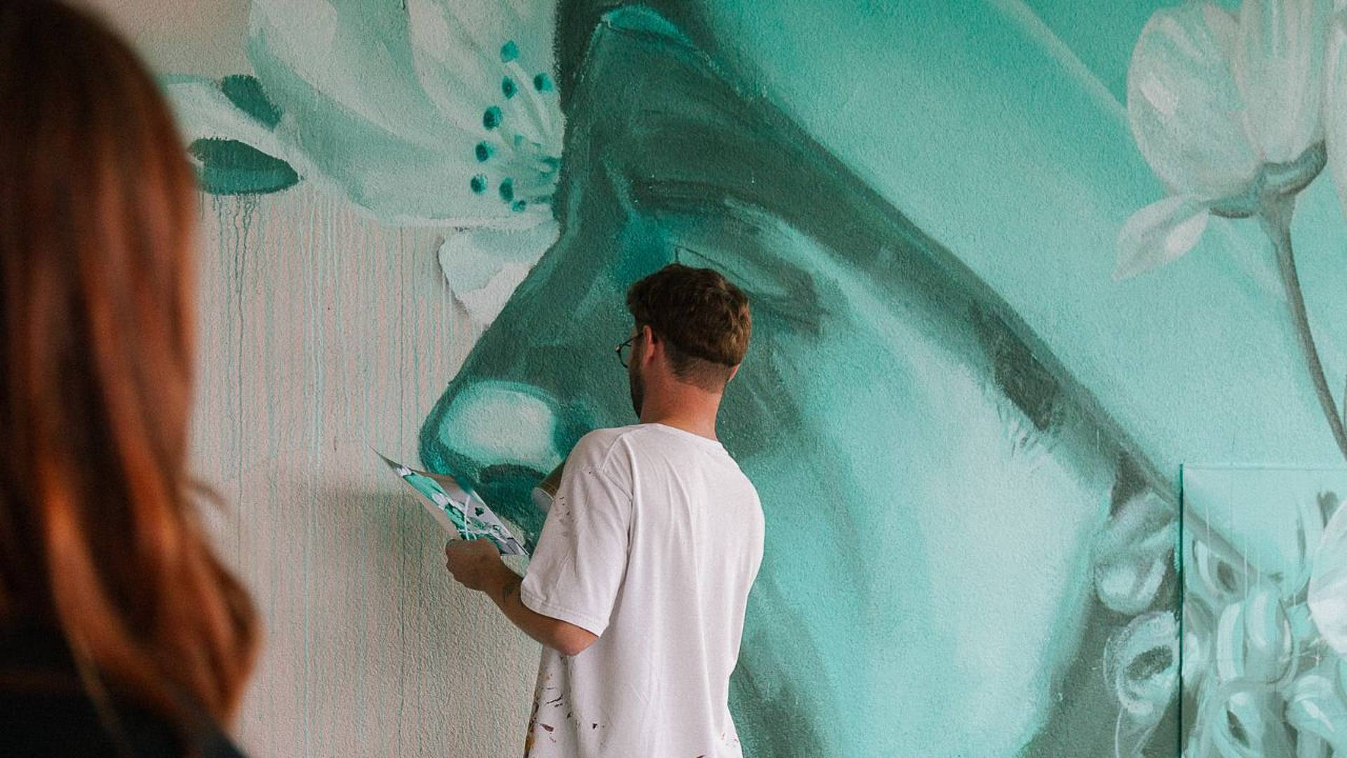 Urban‑Art‑Wandbild: Eine Person arbeitet mit Pinsel und Vorlage an einem grossformatigen Porträt auf einer türkisfarbenen Wand