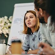 Person im Gespräch während eines Meetings, mit Stift in der Hand und Flipchart im Hintergrund, in einer Büroumgebung.
