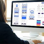 Person arbeitet am Computer an UI/UX-Prototypen mit mobilen App-Layouts und Diagrammen auf dem Bildschirm.