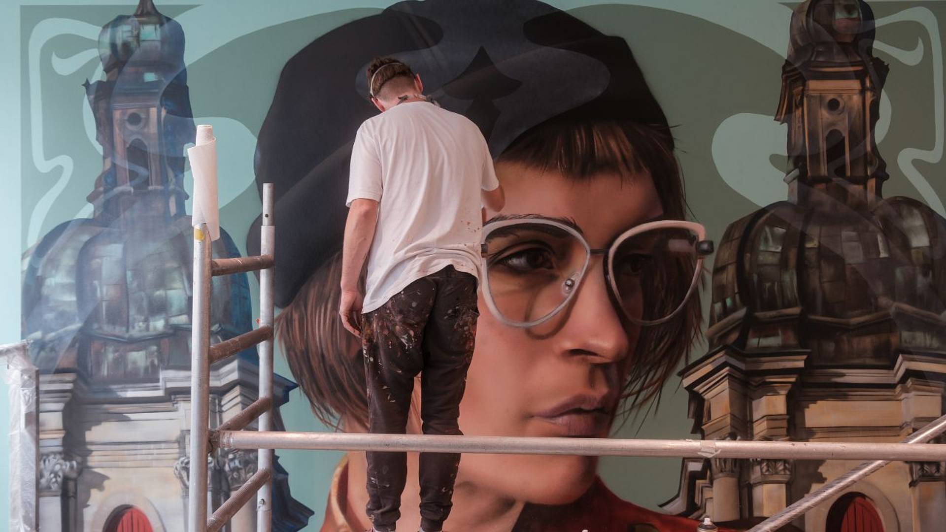 Urban‑Art‑Graffiti: Eine Person steht auf einem Gerüst und malt ein grossformatiges Wandbild mit Porträt und architektonischen Elementen vor hellblauem Hintergrund