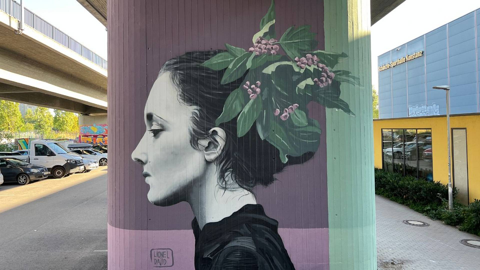 Grossformatiges Urban‑Art‑Porträt im Profil an einer Stützmauer unter einer Brücke, mit gemaltem Kopf und floralen Elementen im Haar vor farbigem Hintergrund