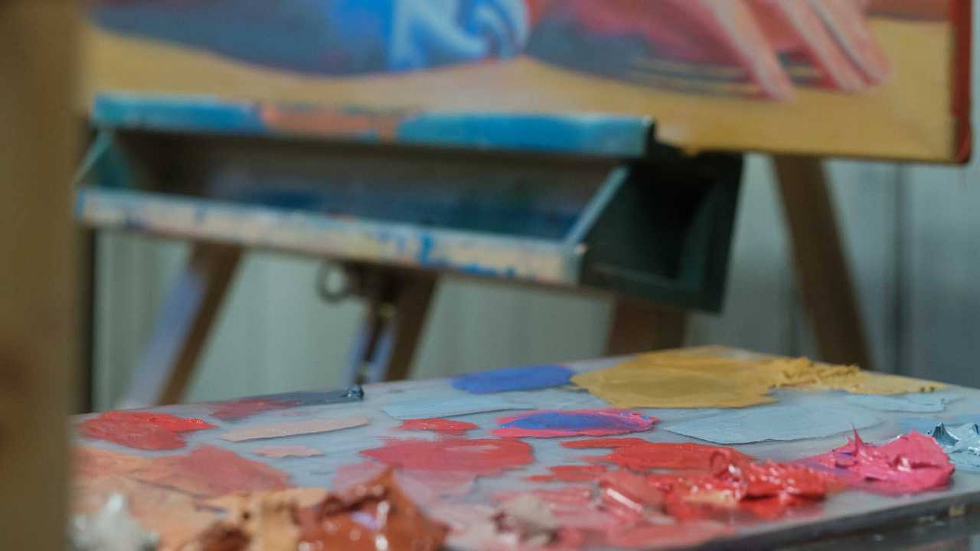 Ölfarben in Rot‑, Blau‑ und Beigetönen auf Malpalette vor einer Staffelei