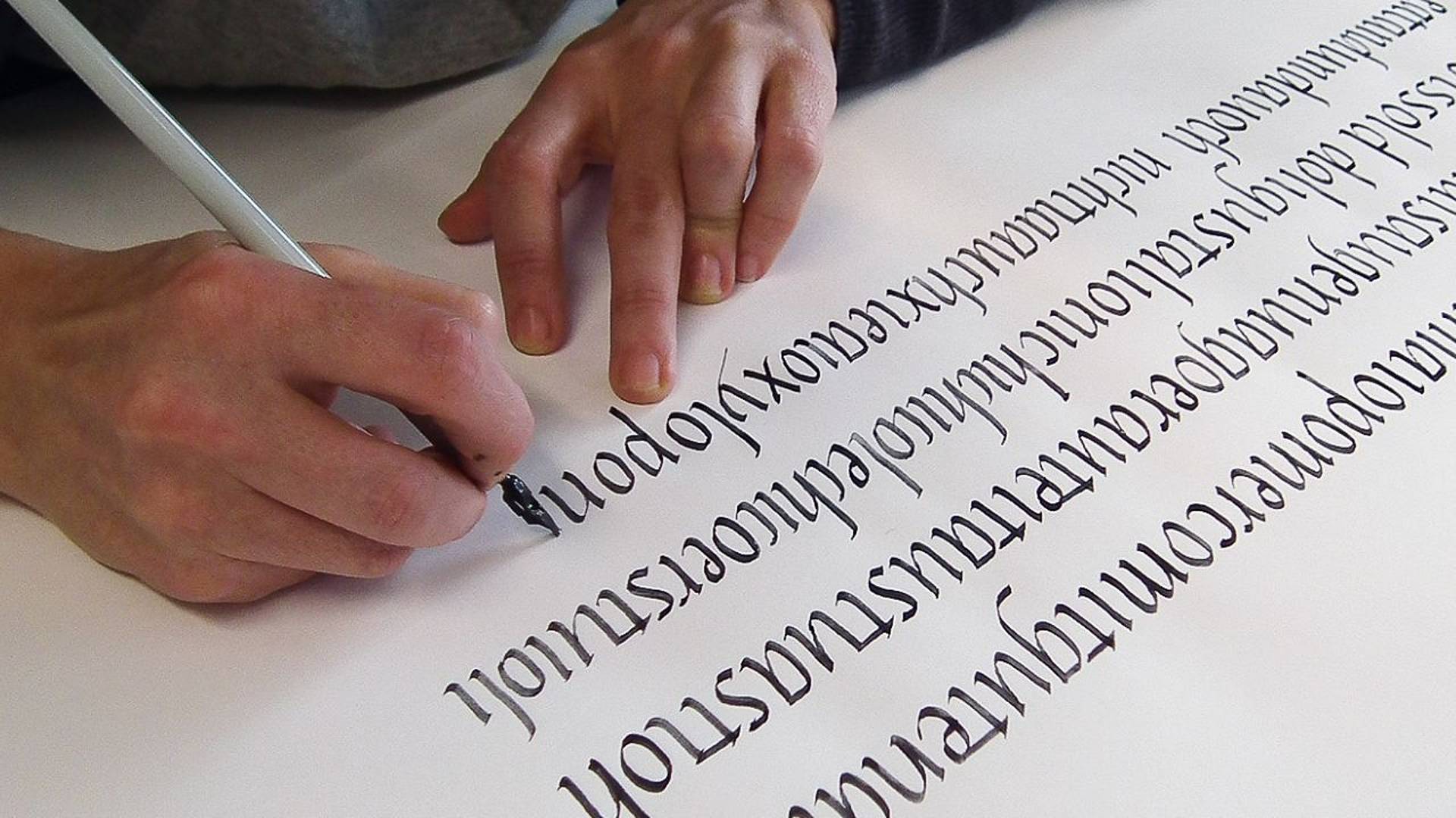 Hände schreiben mit einem schwarzen Kalligrafiestift dekorative Buchstaben auf ein weisses Blatt Papier.