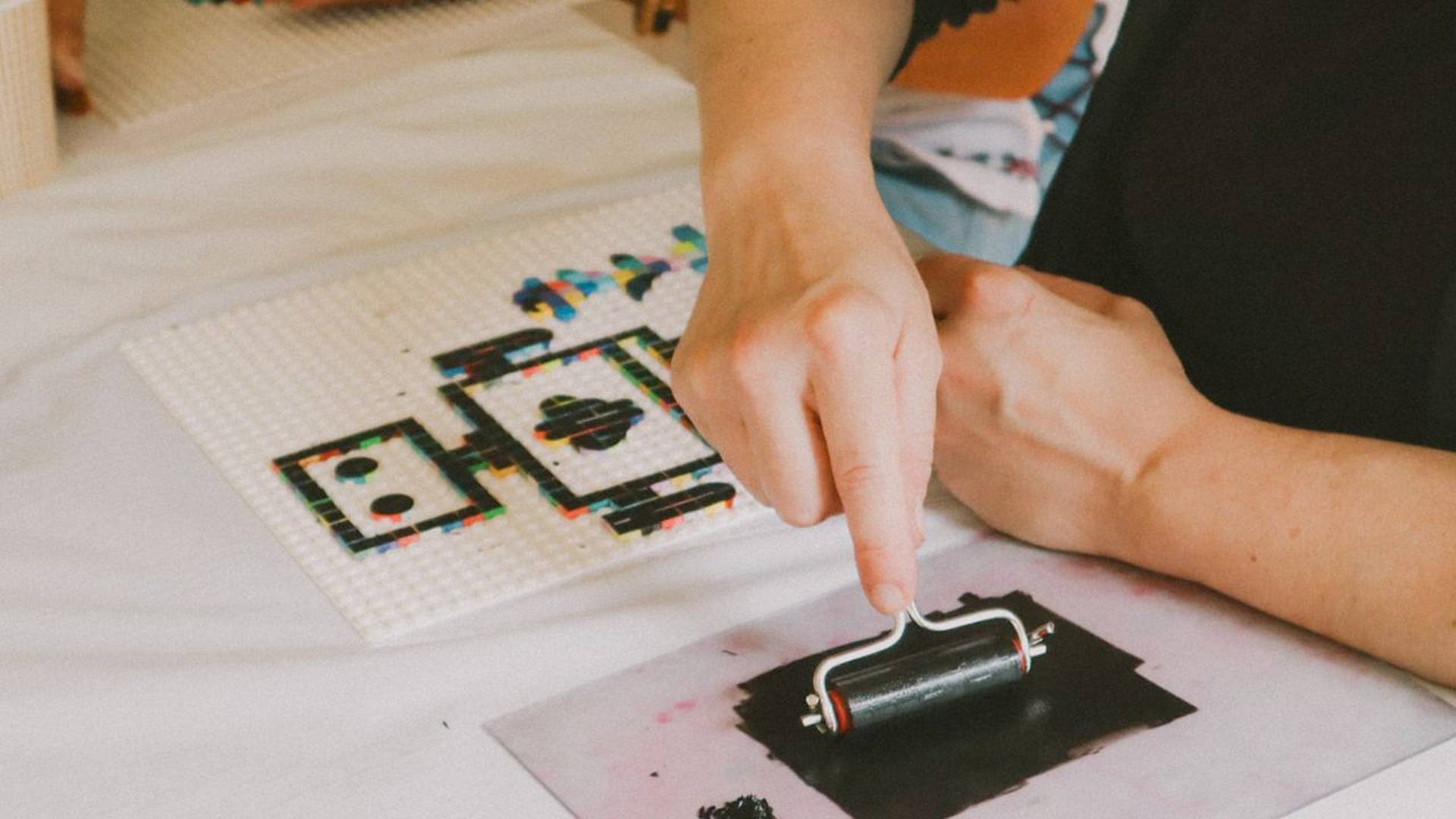 Auftragen von schwarzer Druckfarbe mit einer Farbwalze beim Drucken mit Lego‑Steinen, Lego‑Druckplatte im Hintergrund.