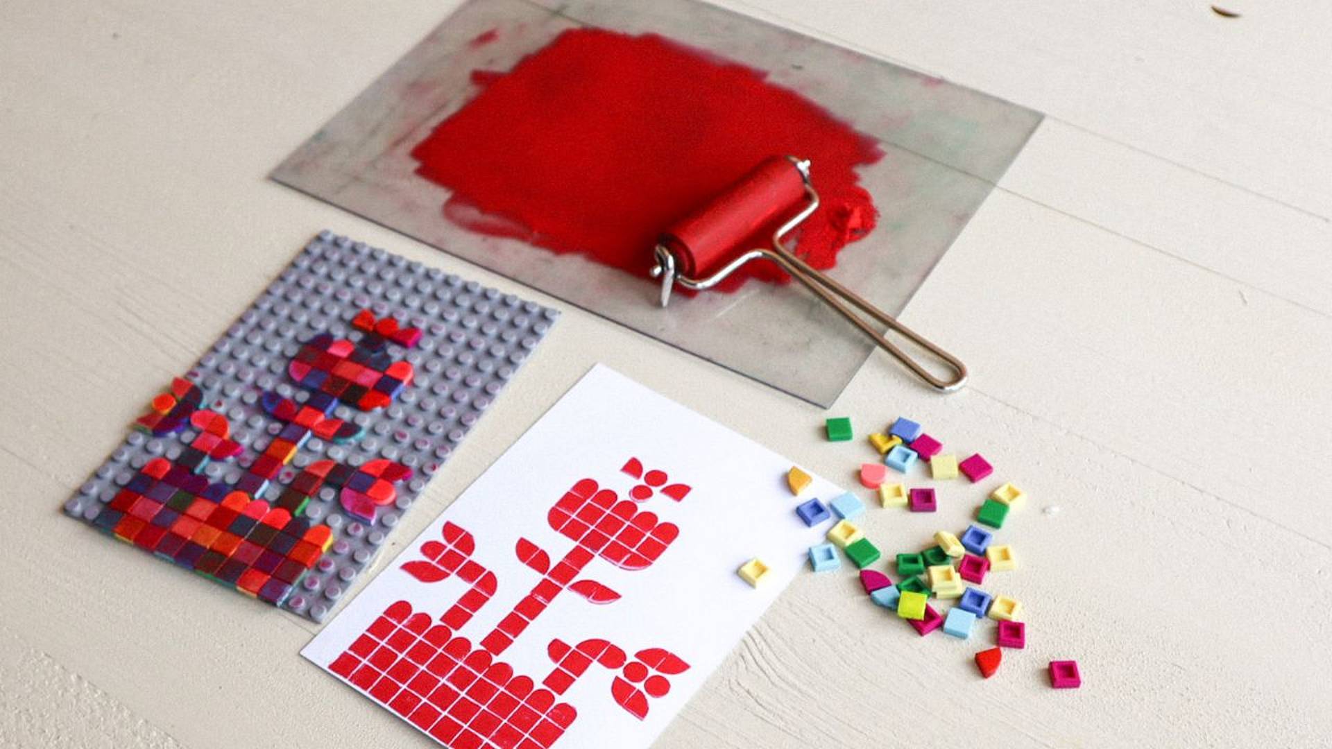 Lego‑Print‑Arbeitsschritt mit Farbwalze, roter Druckfarbe, Lego‑Druckplatte und gedrucktem Motiv auf Papier.