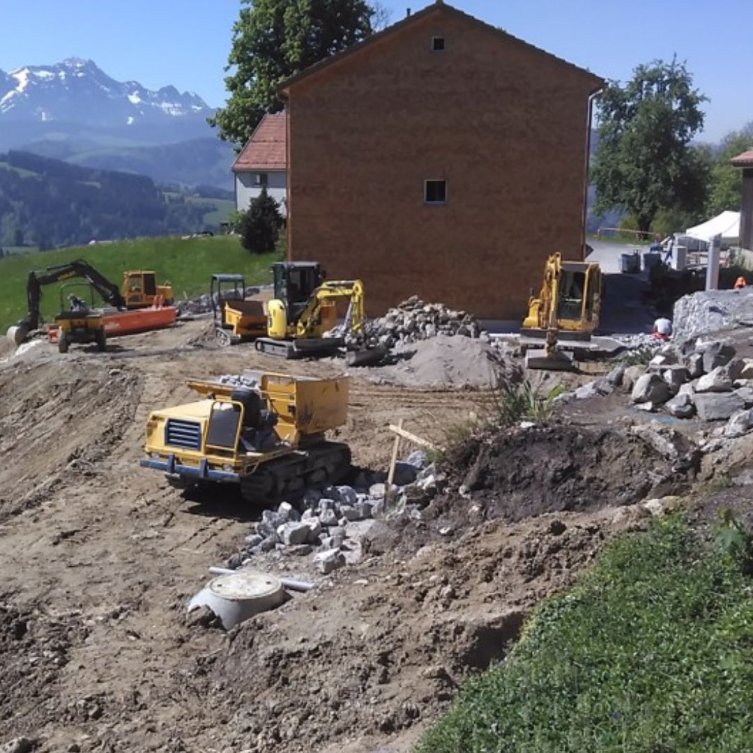 Baustelle im ländlichen Raum mit mehreren Baggern und Erdarbeiten vor alpiner Bergkulisse.