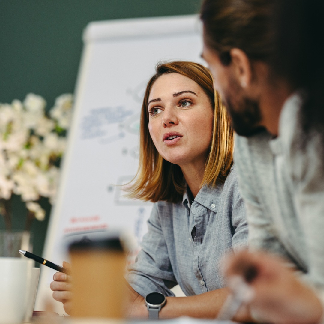 Person im Gespräch während eines Meetings, mit Stift in der Hand und Flipchart im Hintergrund, in einer Büroumgebung.